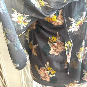 K&ouml;nnte beinhalten: Eine dunkelgraue Bluse mit einem Blumenmuster mit rosa, gelben und wei&szlig;en Blumen. Der transparente Stoff f&auml;llt weich und erzeugt eine flie&szlig;ende Silhouette. Der Blumendruck verleiht dem Kleidungsst&uuml;ck einen Hauch von Eleganz.