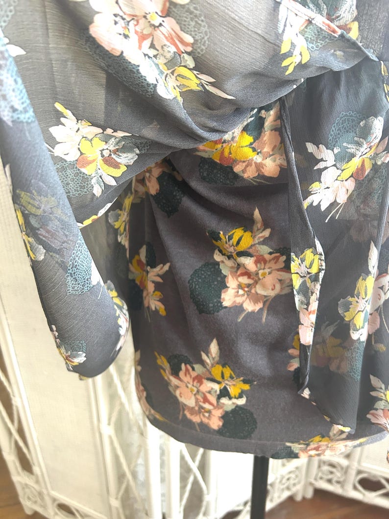 K&ouml;nnte beinhalten: Eine dunkelgraue Bluse mit einem Blumenmuster mit rosa, gelben und wei&szlig;en Blumen. Der transparente Stoff f&auml;llt weich und erzeugt eine flie&szlig;ende Silhouette. Der Blumendruck verleiht dem Kleidungsst&uuml;ck einen Hauch von Eleganz.