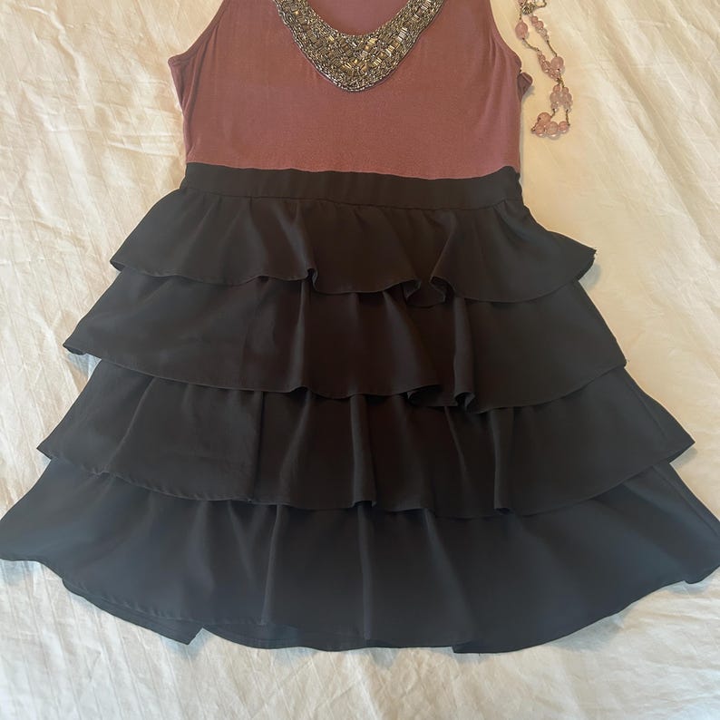 Puede incluir: Un vestido sin mangas con un corpi&ntilde;o malva y una falda negra escalonada. El escote est&aacute; adornado con cuentas plateadas. Un delicado collar rosa est&aacute; cerca. El vestido es adecuado para una ocasi&oacute;n especial.