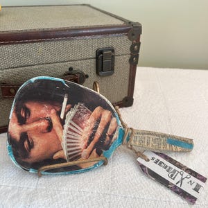 May include: A hand mirror with a collage of a person holding playing cards and a cigarette. The handle is wrapped in newspaper and blue tape. A tag with the text "In Pieces" hangs from the handle. A vintage suitcase is in the background.