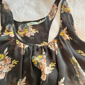 K&ouml;nnte beinhalten: Ein dunkelgraues Tanktop mit einem Blumenmuster in Gelb-, Rosa- und Wei&szlig;t&ouml;nen. Das Top hat ein Racerback-Design und einen Rundhalsausschnitt. Der Stoff scheint durchsichtig zu sein.