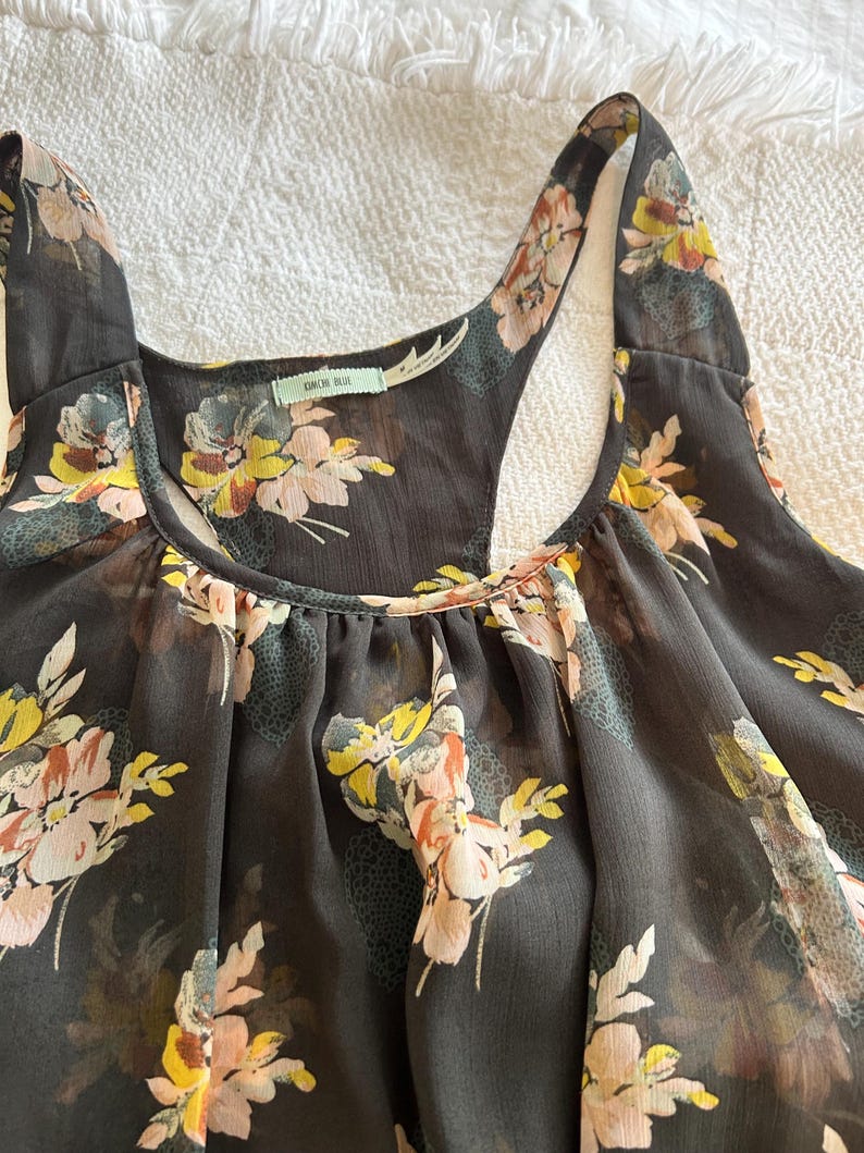 K&ouml;nnte beinhalten: Ein dunkelgraues Tanktop mit einem Blumenmuster in Gelb-, Rosa- und Wei&szlig;t&ouml;nen. Das Top hat ein Racerback-Design und einen Rundhalsausschnitt. Der Stoff scheint durchsichtig zu sein.