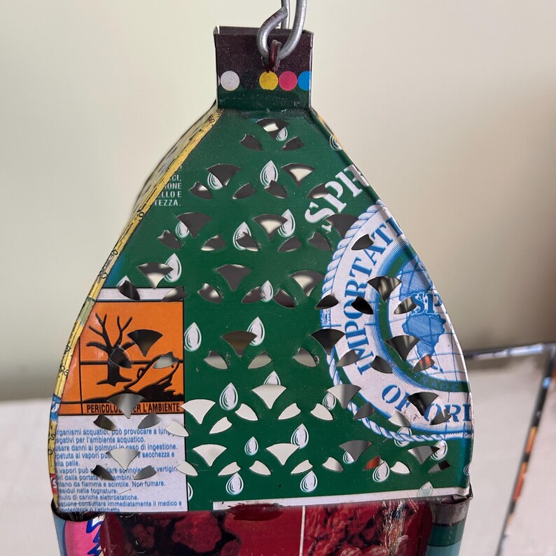 May include: A handmade, decorative lantern crafted from recycled metal. The lantern features a green, white, and red color scheme with cut-out teardrop shapes. The lantern has text and a chain for hanging.