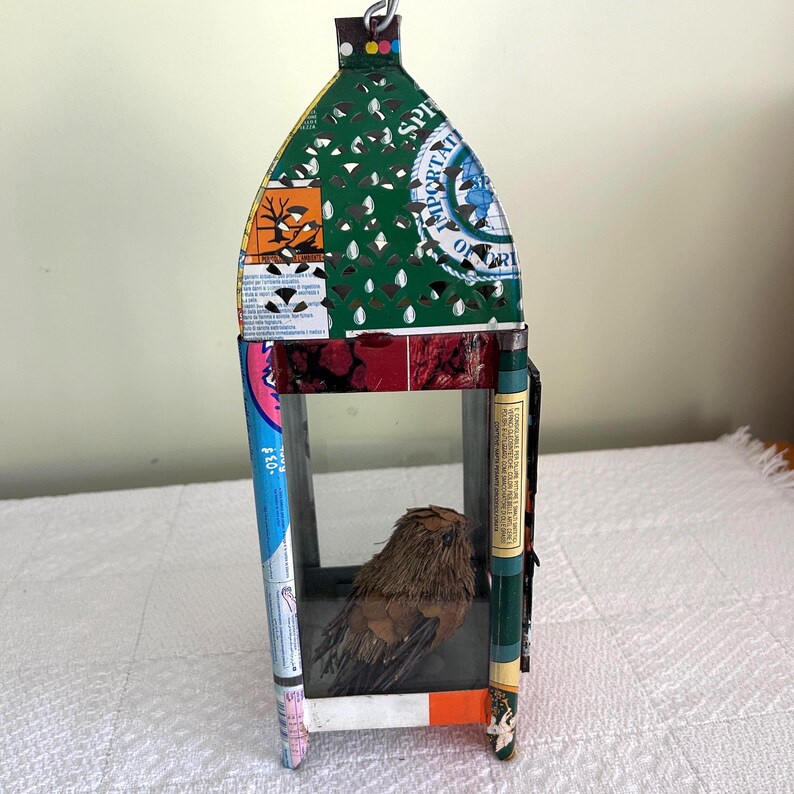 May include: A handmade birdhouse constructed from recycled materials, featuring a small bird figure inside. The birdhouse has a rectangular shape with a peaked roof and is adorned with colorful paper and packaging elements.