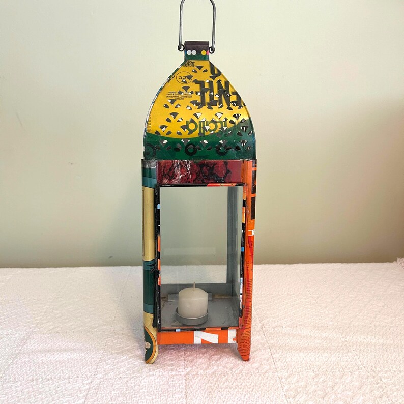 May include: A colorful, handcrafted lantern with a candle inside. The lantern is made from recycled materials, featuring a yellow, green, and orange design. It has a metal handle and a rectangular shape with glass panels.