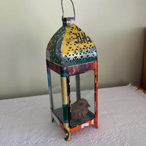 May include: A colorful metal lantern with a handle, featuring a decorative bird inside. The lantern has a rectangular base and a domed top with cut-out patterns. The colors include green, yellow, red, and orange.