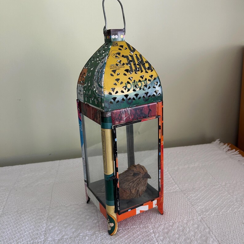 May include: A colorful metal lantern with a handle, featuring a decorative bird inside. The lantern has a rectangular base and a domed top with cut-out patterns. The colors include green, yellow, red, and orange.