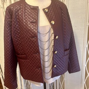 May include: A burgundy quilted jacket with gold buttons and a round neckline. The jacket has a tailored fit with two front pockets and is displayed on a mannequin. The jacket is paired with a white top.