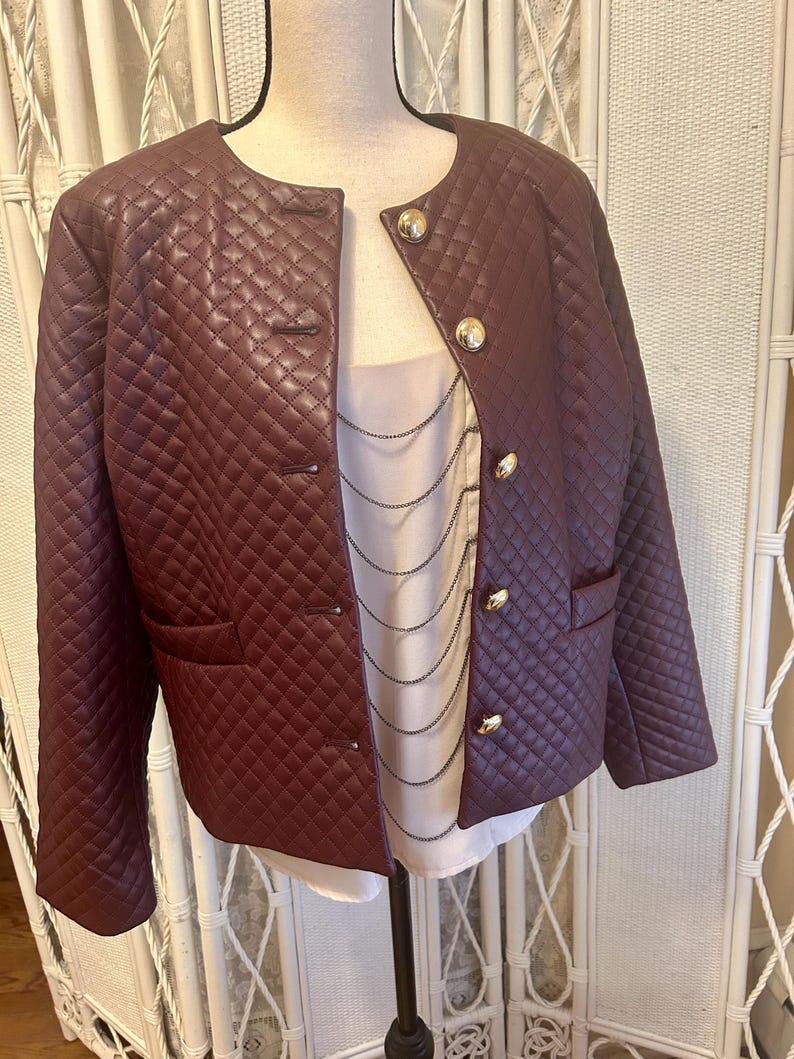 May include: A burgundy quilted jacket with gold buttons and a round neckline. The jacket has a tailored fit with two front pockets and is displayed on a mannequin. The jacket is paired with a white top.