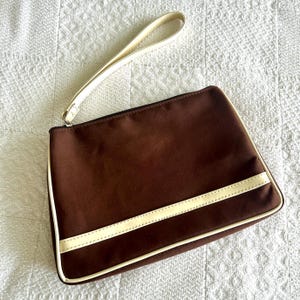 Op de afbeelding: Een bruine clutch met een crèmekleurige polsband en een crèmekleurige streep over de voorkant. De clutch heeft een ritssluiting en crèmekleurige rand. De tas ligt op een witte, getextureerde ondergrond.