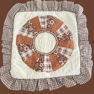 May include: Square pillow with a white center and a circular patchwork design in shades of brown, orange, and white. The pillow has a ruffled border in a brown and white gingham pattern.