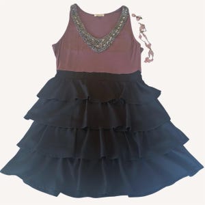 Puede incluir: Un vestido sin mangas con un corpi&ntilde;o malva y una falda negra escalonada. El escote est&aacute; adornado con cuentas plateadas. Un collar de cuentas a juego est&aacute; drapeado cerca. El vestido es una prenda elegante.