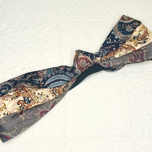 May include: A patterned headband featuring a paisley design in navy blue, cream, burgundy, and gray. The fabric is twisted at the center, creating a stylish accessory. The headband is designed to be worn around the head.
