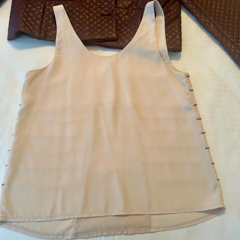 May include: A light peach-colored tank top with a V-neck design. The tank top has horizontal lines across the front and small metal accents along the sides. A burgundy quilted jacket is visible in the background.