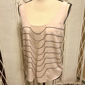 May include: A light pink sleeveless top with a scoop neckline and a series of wavy, dark gray beaded embellishments. The top is made of a silky material and is displayed on a mannequin.