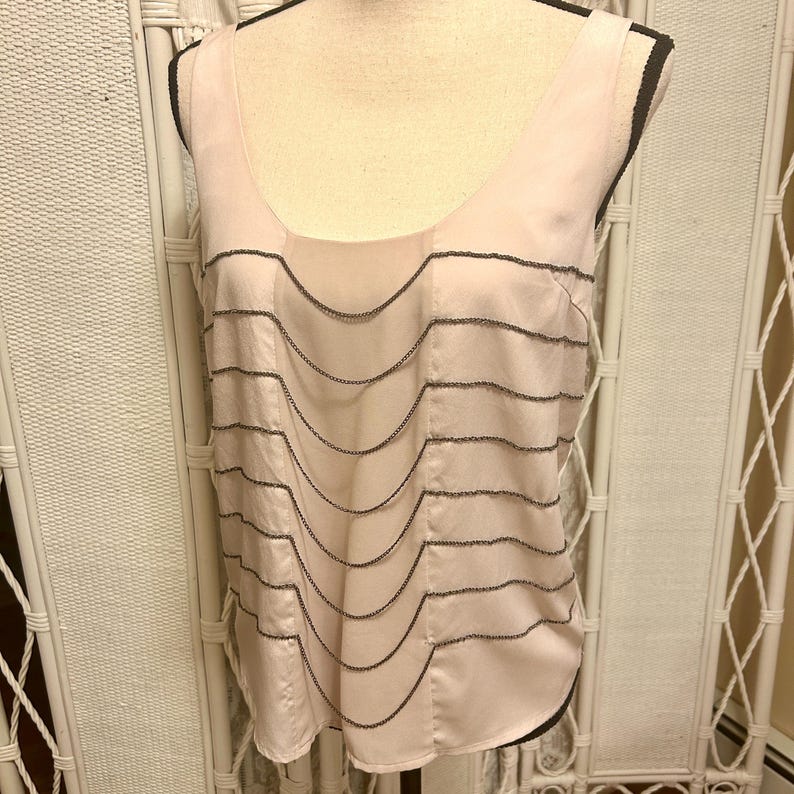 May include: A light pink sleeveless top with a scoop neckline and a series of wavy, dark gray beaded embellishments. The top is made of a silky material and is displayed on a mannequin.