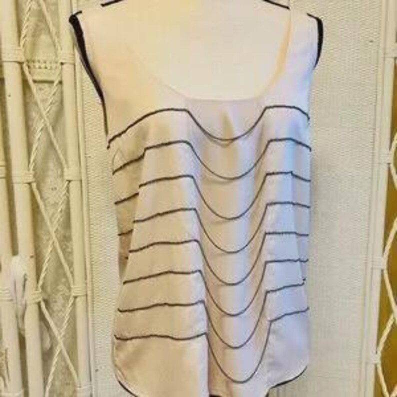 May include: A sleeveless, light pink top with a scoop neck and a layered design. The layers are outlined with a dark gray trim. The top is displayed on a mannequin.