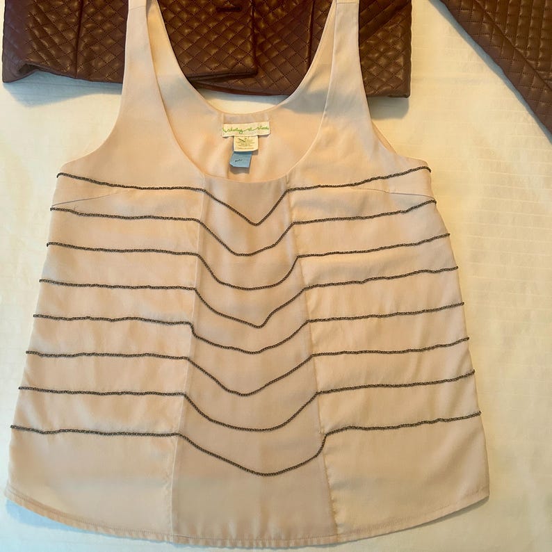 May include: A light pink sleeveless top with horizontal beaded lines and a scoop neckline. The top has a flowing design and a tag near the neckline. A brown quilted jacket is visible in the background.