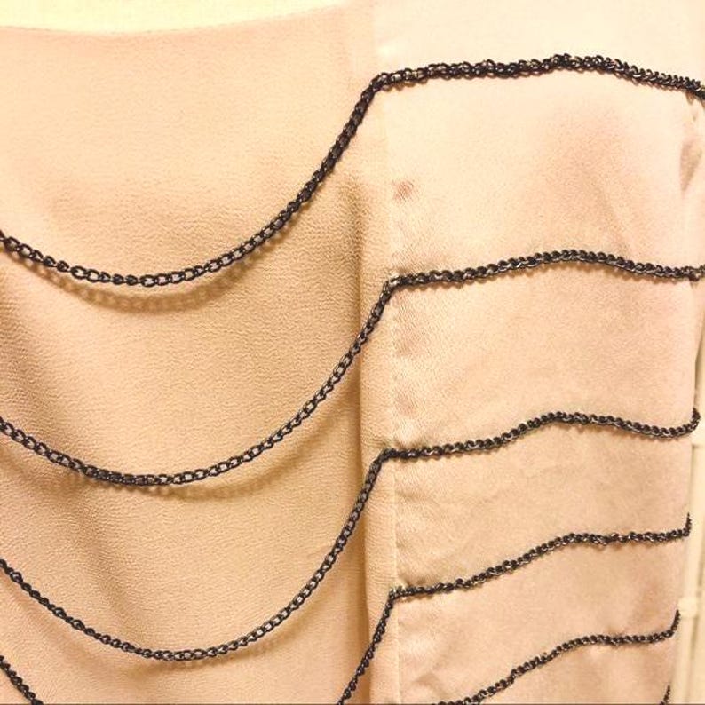 May include: A close-up of a light peach-colored garment with multiple strands of dark gray chain detailing. The chains are arranged in a wavy, layered pattern across the fabric, creating a textured effect. The fabric appears to be a soft, flowing material.