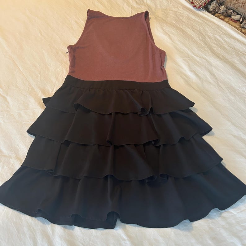 Puede incluir: Vestido sin mangas con corpi&ntilde;o malva y falda negra con volantes escalonados. El vestido est&aacute; tendido sobre una superficie blanca, mostrando su dise&ntilde;o. El vestido parece estar hecho de una tela suave y fluida.
