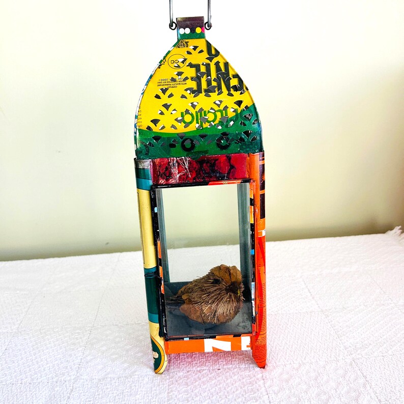 May include: A colorful, handmade lantern with a bird figurine inside. The lantern is constructed from recycled materials, featuring a yellow top with cut-out designs, green and red accents, and an orange and blue frame. A small, brown bird sits inside.