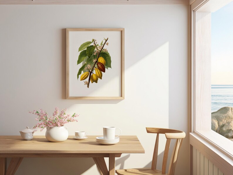 May include: A framed botanical print of a cocoa plant branch with green leaves and yellow and brown cocoa pods. The print is in a light wood frame and hangs on a white wall. A wooden table with a white vase and tea set is in the foreground.