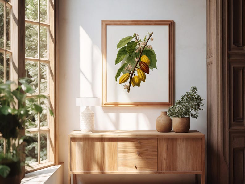 May include: A framed botanical print of a cacao branch with green leaves and yellow and brown pods. The print is displayed on a wooden cabinet with a lamp and potted plants. The room has a window with natural light.