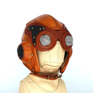 May include: A brown leather aviator helmet with a black visor and mesh goggles. The helmet has a stitched design and is on a white mannequin head.