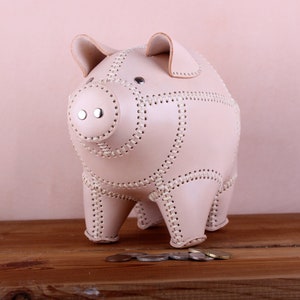 May include: A light pink leather piggy bank with stitched detailing and a coin slot on the top. The piggy bank is standing on a wooden surface with several coins in front of it.