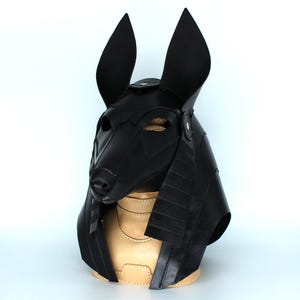 May include: A black leather Anubis mask with a detailed design. The mask has large ears and a pointed snout. The mask is on a tan leather stand.