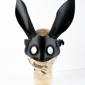 Peut inclure: Masque de lapin en cuir noir avec de grandes oreilles et des trous ronds pour les yeux. Le masque est sur une t&ecirc;te de mannequin.