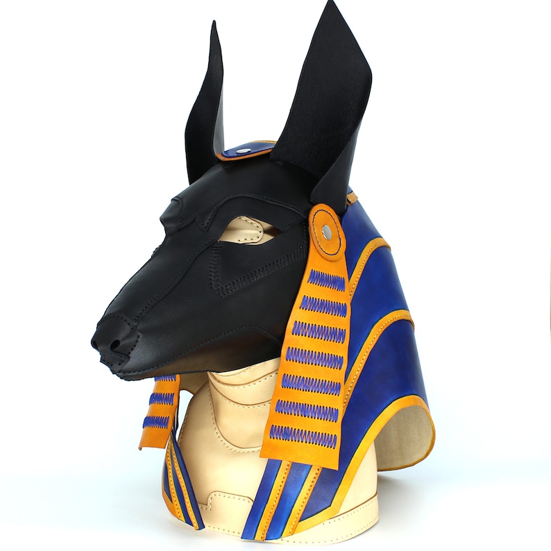 May include: A black leather Anubis mask with blue and yellow accents. The mask has a pointed snout and large ears. The mask is decorated with a blue and yellow patterned fabric.