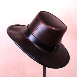 May include: A brown leather cowboy hat with a black band and stitching detail. The hat is on a stand.