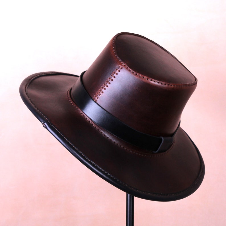 May include: A brown leather cowboy hat with a black band and stitching detail. The hat is on a stand.