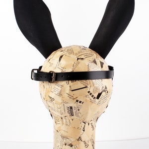 Peut inclure: Un masque de lapin noir avec de longues oreilles et une sangle en cuir noir. Le masque est en papier m&acirc;ch&eacute; et recouvert de coupures de journaux.