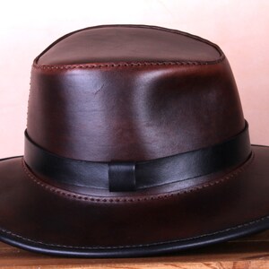 May include: A brown leather fedora hat with a black leather band. The hat has a wide brim and a stitched edge.