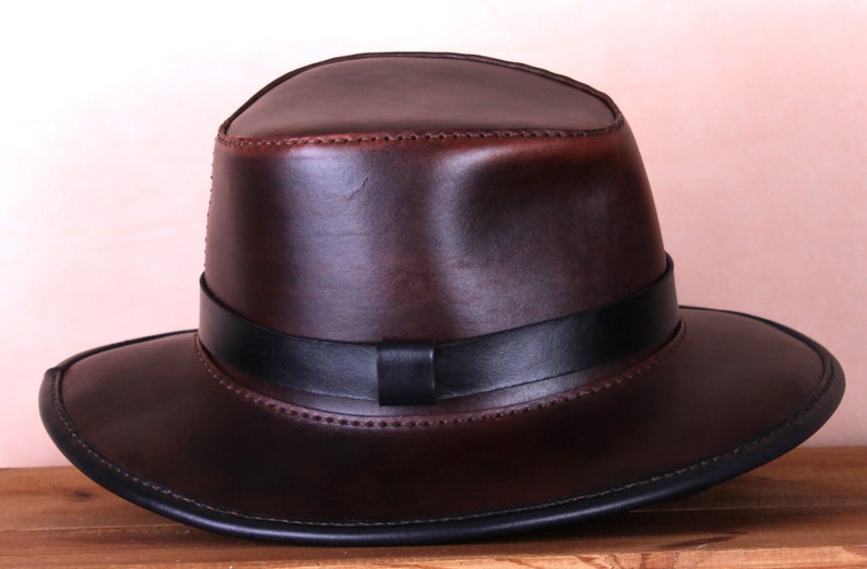 May include: A brown leather fedora hat with a black leather band. The hat has a wide brim and a stitched edge.