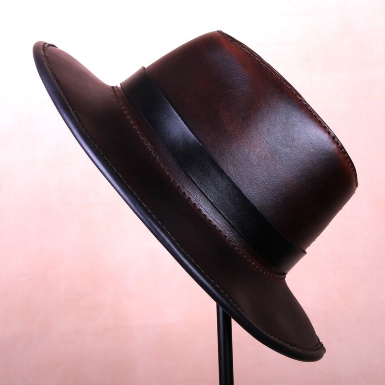 May include: A brown leather fedora hat with a black band. The hat is on a stand and has a slightly distressed look.