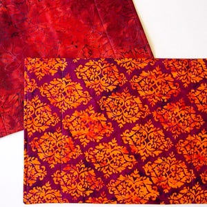 May include: Two rectangular placemats. One is a deep red with a floral pattern. The other is a deep purple with an orange and gold damask design. The placemats are made of fabric and have a quilted appearance.