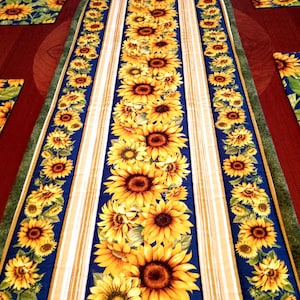 May include: A long rectangular table runner featuring a central panel of vibrant yellow sunflowers on a deep blue background, flanked by vertical stripes of tan and cream, with matching sunflower patterned placemats visible on the sides.