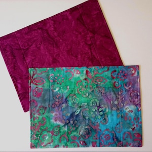 May include: Two rectangular placemats. One is a deep magenta with a mottled texture. The other is a vibrant mix of teal, green, and purple with a floral batik pattern. Both are quilted.