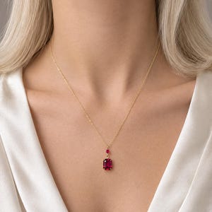 May include: A gold chain necklace featuring a rectangular, deep red gemstone pendant suspended from a smaller, round red stone accent. The pendant is set in a gold-toned metal frame, creating a delicate piece of jewellery that rests against the collarbone.