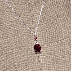 May include: A silver necklace with a rectangular, deep red pendant. The pendant is accented with a smaller, round red stone above it. The necklace hangs on a silver chain, creating a classic and elegant look.