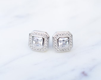 May include: A pair of silver-toned stud earrings. Each earring features a square, clear gemstone center, surrounded by a halo of smaller, clear stones. The earrings are set on a white surface.