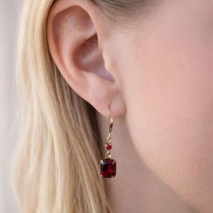 May include: A gold-toned leverback earring featuring a small round red gemstone suspended above a larger rectangular red gemstone. The earring is secured to the earlobe with a thin gold-toned wire hook, showcasing a classic drop design.