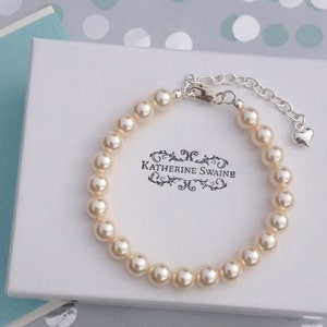 Puede incluir: Un brazalete de perlas blancas con una cadena de plata y un dije en forma de corazón. El brazalete está en una caja blanca con el texto "Katherine Swaine" impreso en ella.