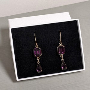 May include: A pair of dangle earrings with a gold setting and two purple gemstones. The larger gemstone is a square shape and the smaller gemstone is a teardrop shape.
