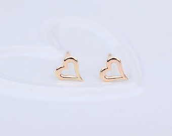 Puede incluir: Un par de pendientes de botón dorados con forma de corazón. Los pendientes tienen un contorno delicado en forma de corazón. Se exhiben sobre una superficie blanca, creando una presentación limpia y elegante. Un accesorio romántico.