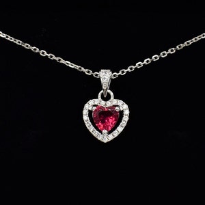 May include: A silver chain necklace with a heart-shaped pendant. The pendant features a red gemstone surrounded by smaller clear gemstones.