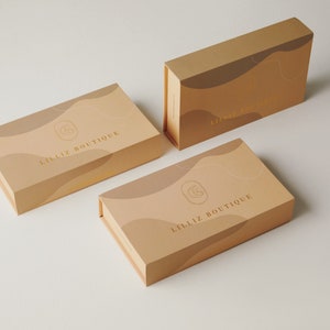 Op de afbeelding: Drie dozen met een beige achtergrond en gouden accenten. De dozen hebben een golvend ontwerp en de tekst "LILLIZ BOUTIQUE" staat op de voorkant.